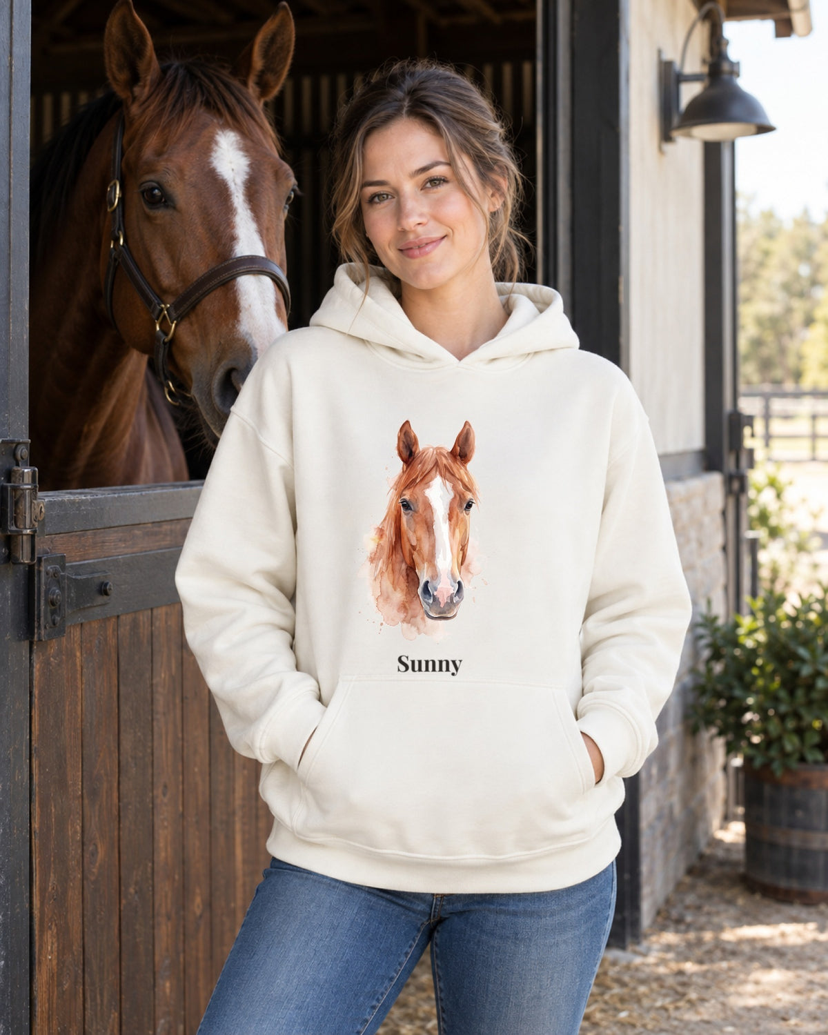 Hoodie in Creme mit Portrait von einem braunen Pferd im Stil Aquarell und dem Namen Sunny, getragen von einer Frau mit ihrem Pferd am Stall