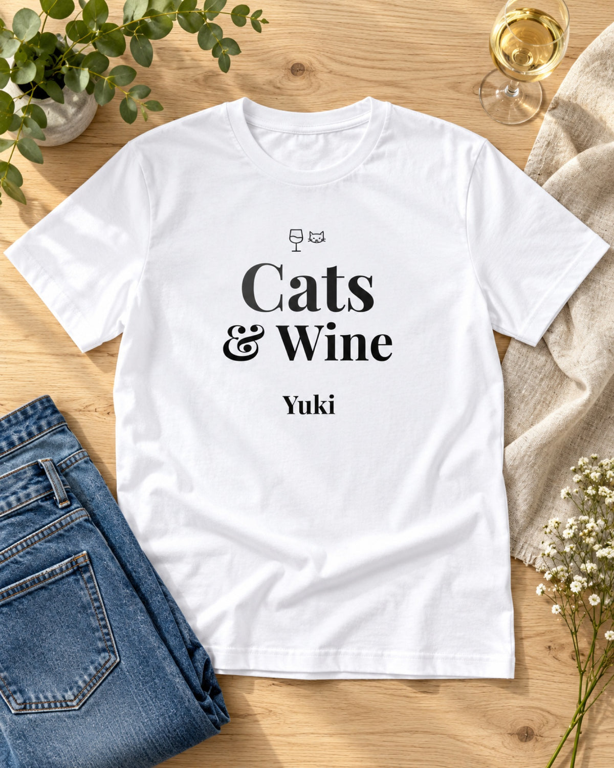 Weißes T-Shirt Cats und Wine mit Namen Yuki, flach gelegt auf Holz mit Jeans, Weinglas und Blumen