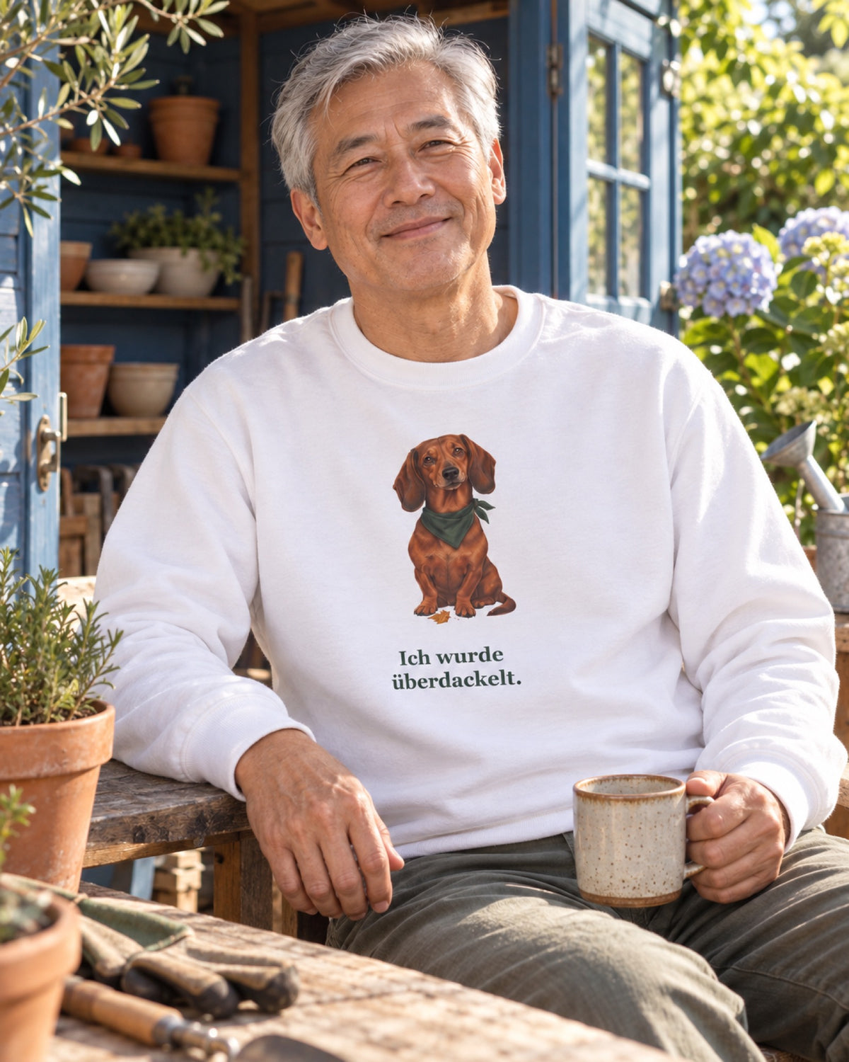 Mann im weißen Sweatshirt mit festem Dackelmotiv und dem Aufdruck Ich wurde überdackelt vor einem blauen Gartenhaus