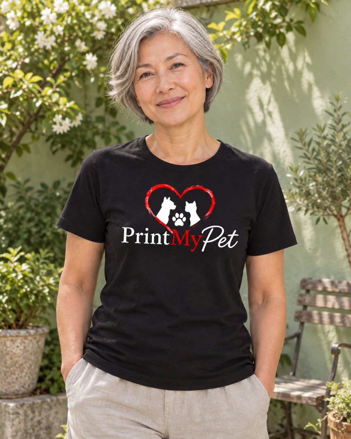 Frau im schwarzen PrintMyPet T-Shirt mit großem Logo auf der Brust vor einer hellgrünen Sommergartenmauer