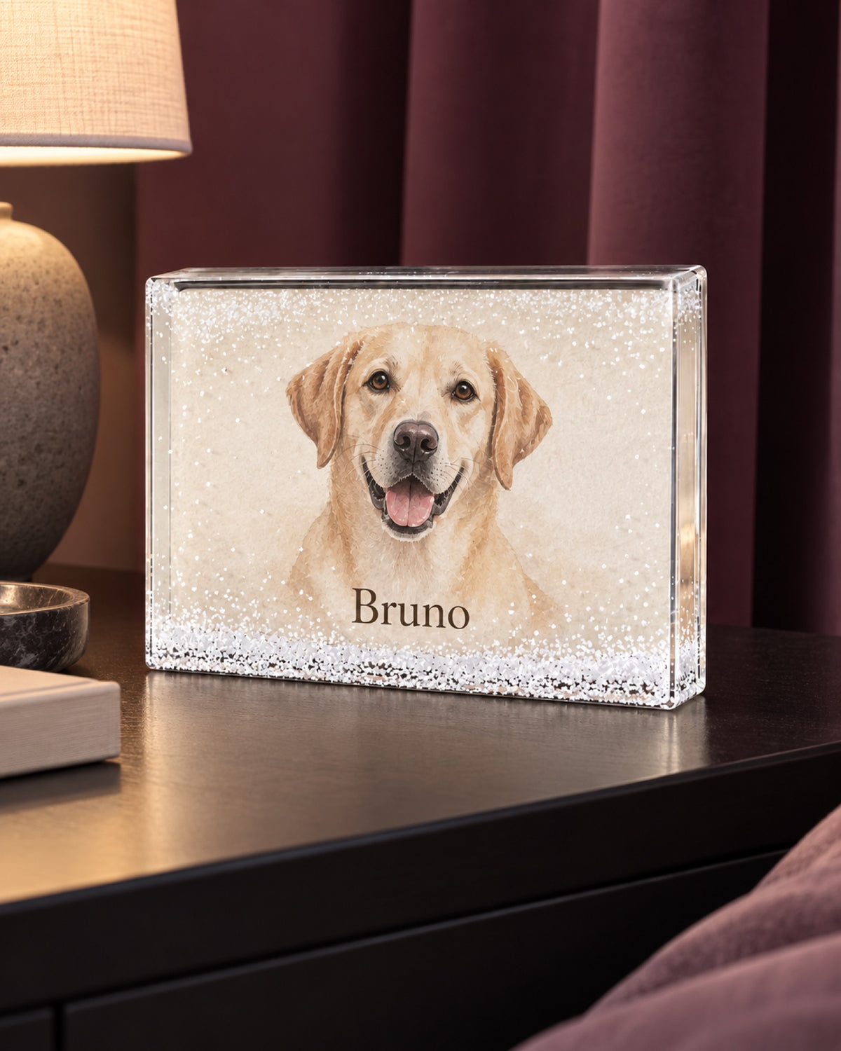 Schüttelrahmen mit weißen Schneeflocken und gelber Labrador Bruno auf einem schwarzen Nachttisch vor pflaumenfarbenen Vorhängen