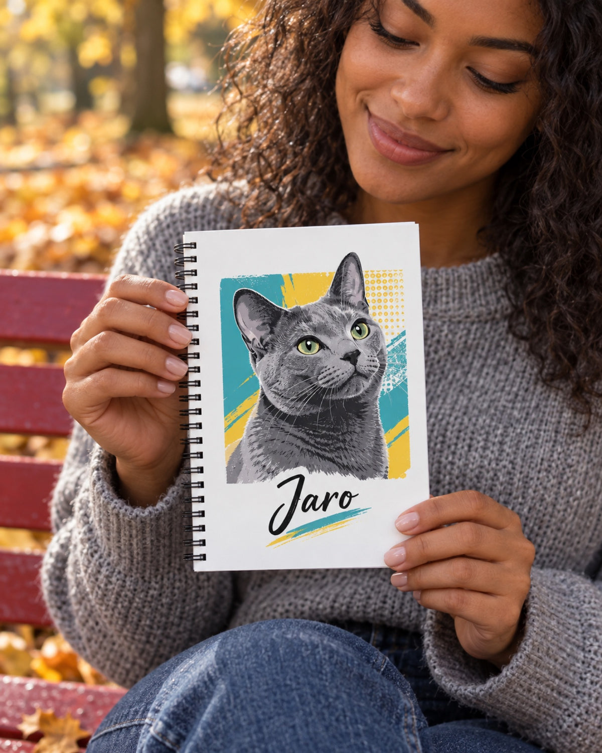Spiralnotizbuch 14 × 22 cm mit grauer Katze im Pop Art Stil und Namen Jaro auf den Händen einer jungen Frau mit dunkler Haut und grauem Pullover auf einer roten Parkbank im Herbst
